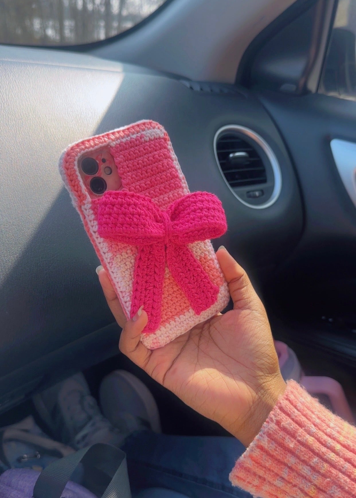 Hand holding a pink crocheted phone case with a bow in a car.