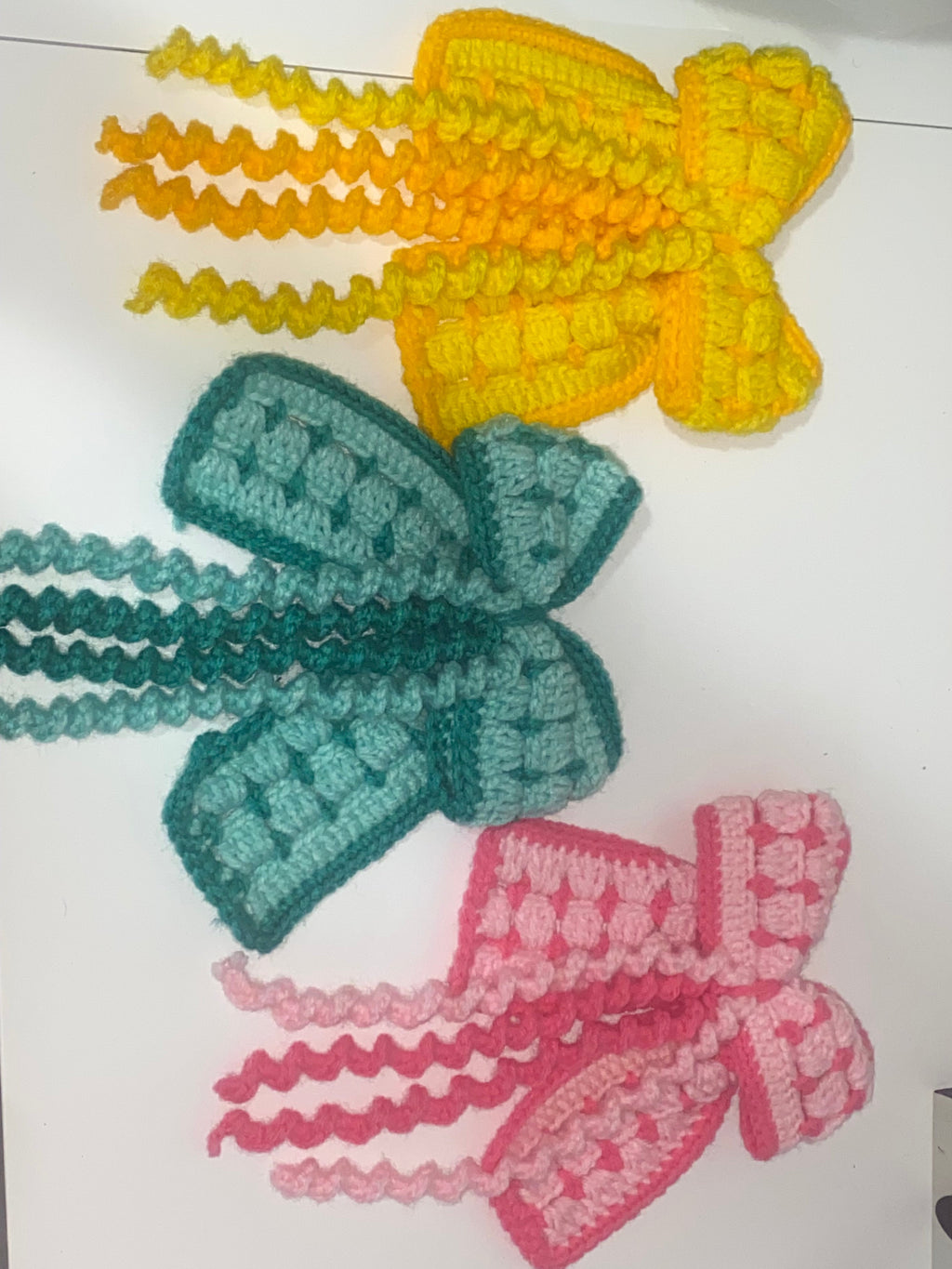Three crocheted bows in yellow, green, and pink on a white surface.